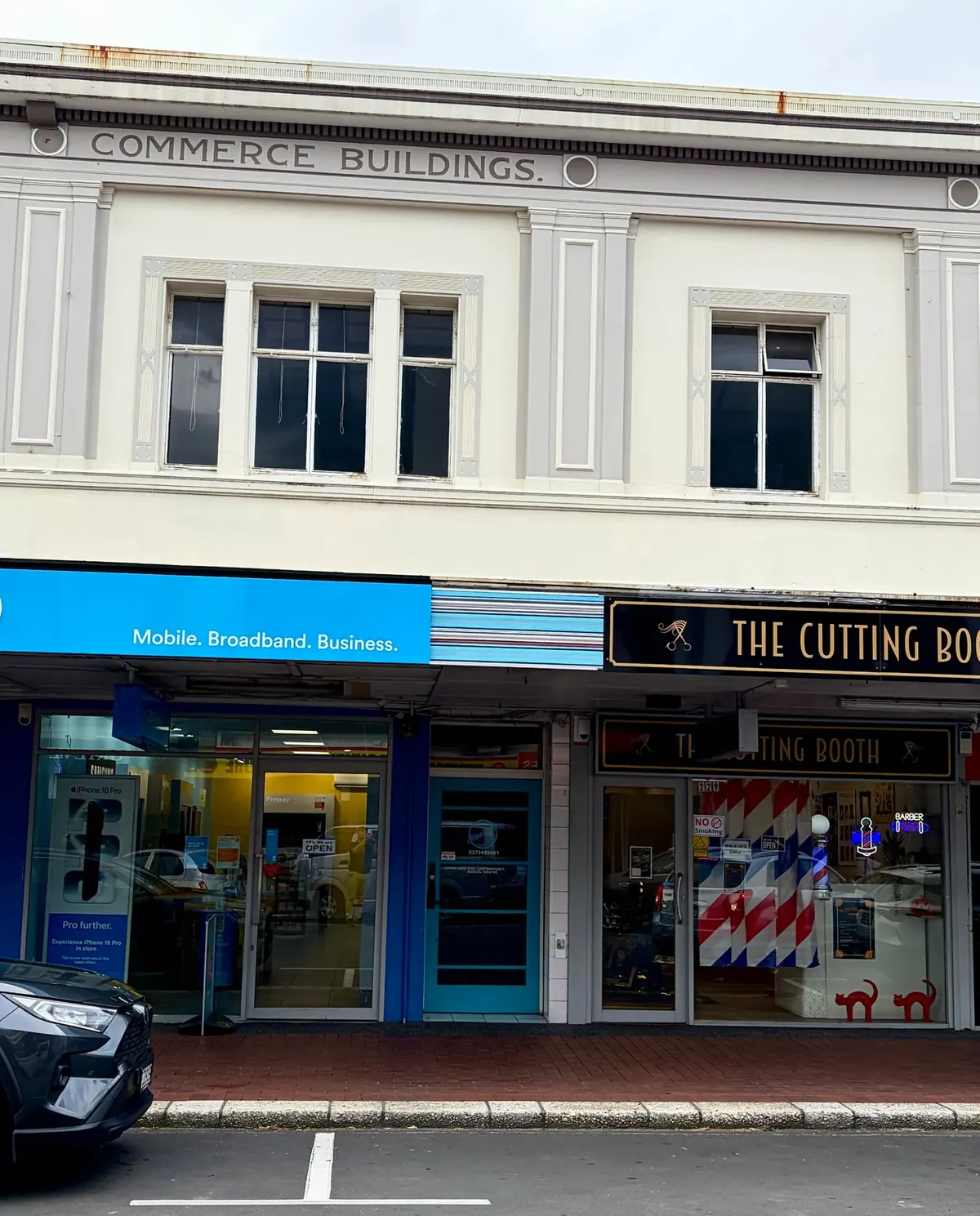 A Strip Painting of sky blue, navy, blush and cream bands on the timber fascia at 227 Heretaunga Street West, between the blue 2degrees sign and the black The Cutting Booth sign, under the Commerce Buildings facade