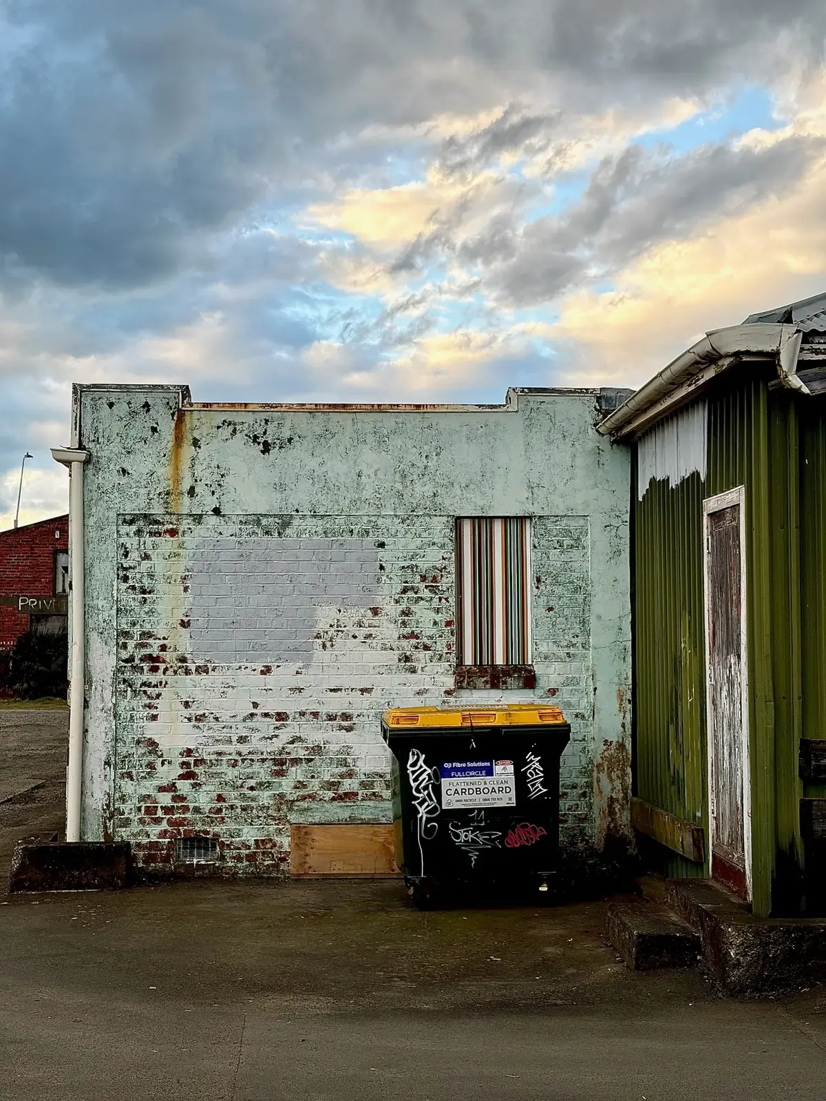 An upright Strip Painting of cream, brick red, maroon, green and orange bands filling the boarded window of a mint-painted brick gable in the 300 West laneway at dusk